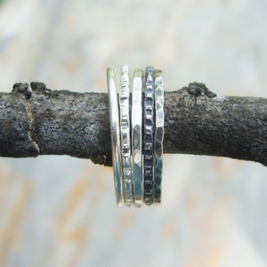 May include: Five silver stacking rings with different textures, including hammered and patterned, are displayed on a branch.