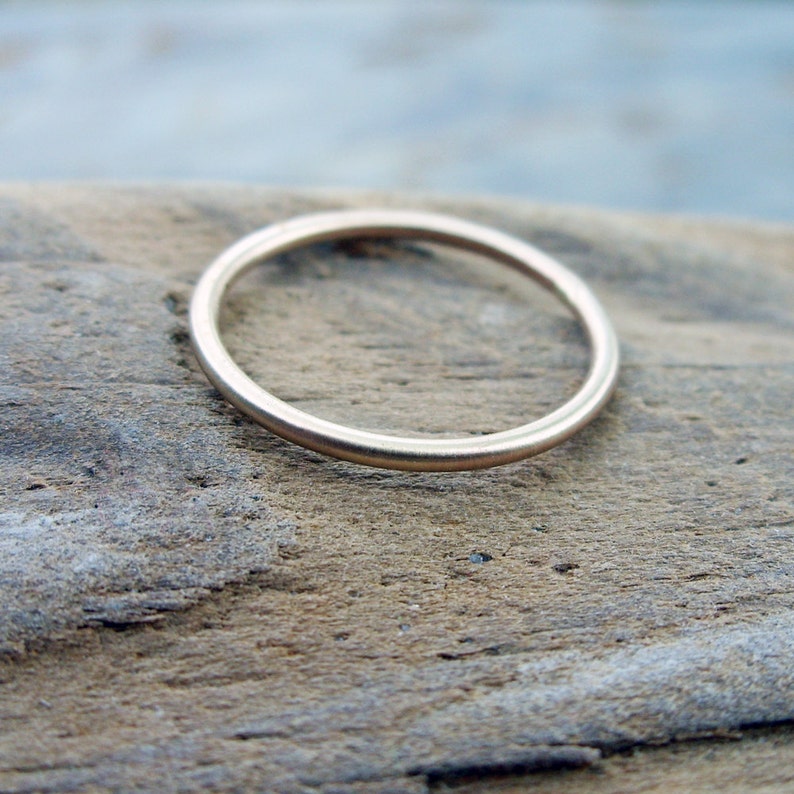 Op de afbeelding: Een dunne, goudkleurige ring. De ring is cirkelvormig en lijkt van metaal te zijn. De ring ligt op een stuk verweerd hout. De ring is eenvoudig en elegant, geschikt voor dagelijks gebruik.