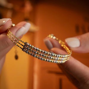 Könnte beinhalten: Ein goldenes Armband mit drei Strängen aus kleinen, strukturierten Perlen. Das Armband wird vor einem unscharfen, orangefarbenen Hintergrund gehalten. Das Armband hat ein filigranes Design und einen Verschluss.