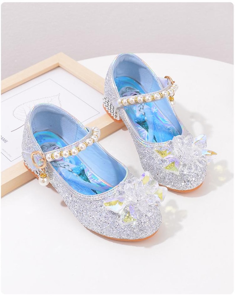 Puede incluir: Zapatos Mary Jane plateados con purpurina, con una correa adornada con perlas y una flor transparente con forma de gema en la puntera. Los zapatos tienen un interior azul claro y un tac&oacute;n peque&ntilde;o.