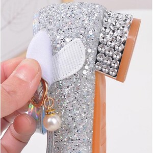 Puede incluir: Zapato plateado con purpurina, lazo blanco y un charm de perla. El zapato tiene suela marr&oacute;n y est&aacute; decorado con pedrer&iacute;a. El texto "Convenient Velcro design" es visible.