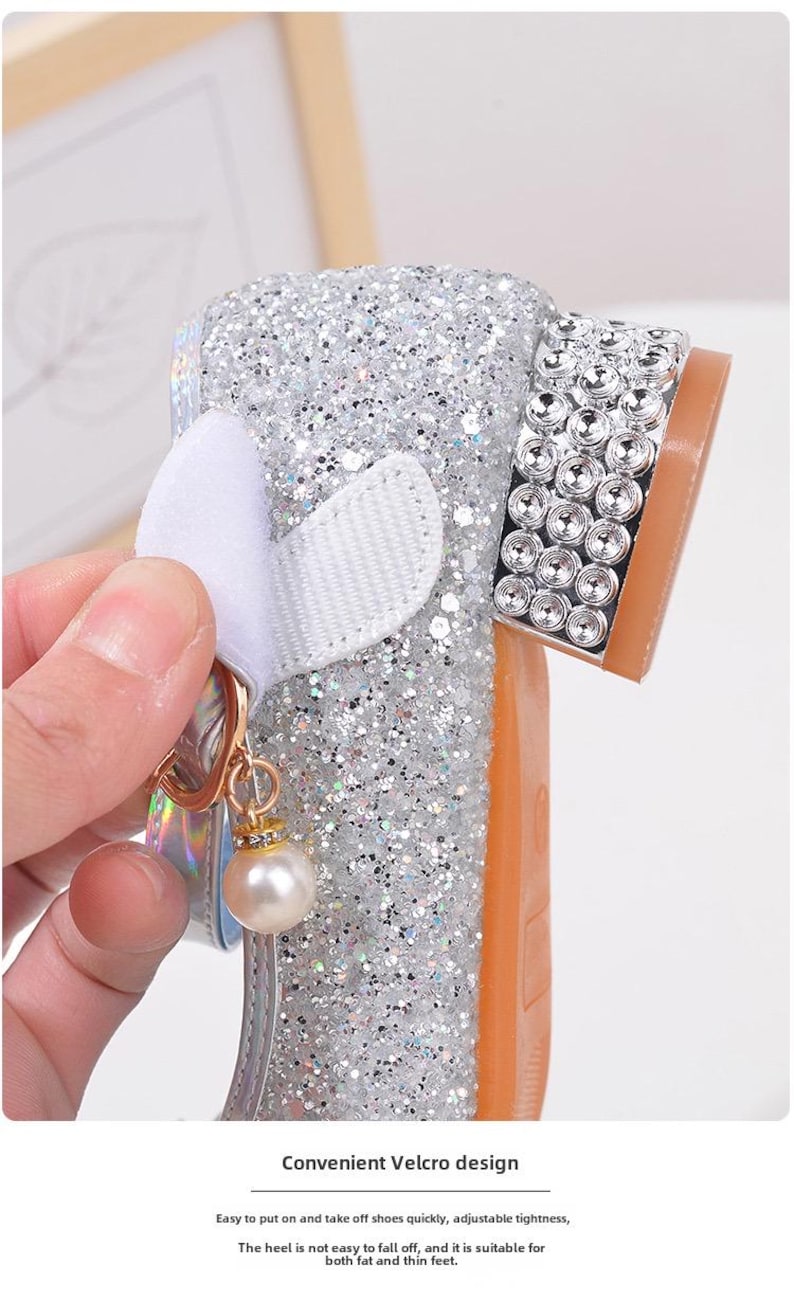 Puede incluir: Zapato plateado con purpurina, lazo blanco y un charm de perla. El zapato tiene suela marr&oacute;n y est&aacute; decorado con pedrer&iacute;a. El texto "Convenient Velcro design" es visible.