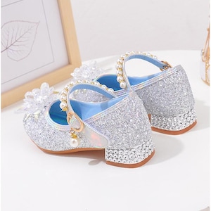 Puede incluir: Zapatos plateados de estilo Mary Jane con purpurina y tac&oacute;n de bloque peque&ntilde;o. Los zapatos tienen una correa con hebilla dorada y perlas, y est&aacute;n adornados con perlas y detalles florales transparentes. Los tacones est&aacute;n adornados con peque&ntilde;os diamantes de imitaci&oacute;n transparentes.