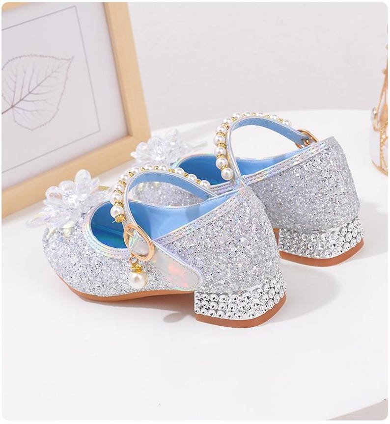 Puede incluir: Zapatos plateados de estilo Mary Jane con purpurina y tac&oacute;n de bloque peque&ntilde;o. Los zapatos tienen una correa con hebilla dorada y perlas, y est&aacute;n adornados con perlas y detalles florales transparentes. Los tacones est&aacute;n adornados con peque&ntilde;os diamantes de imitaci&oacute;n transparentes.