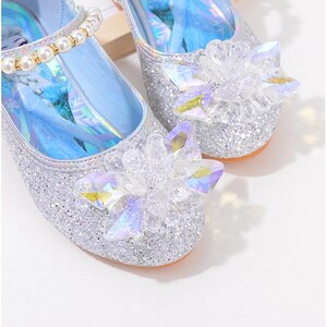 Puede incluir: Zapatos plateados de tela de lentejuelas con adornos de flores de cristal. Los zapatos tienen una correa adornada con perlas y un interior azul claro. El texto de la imagen dice "sequined fabric".