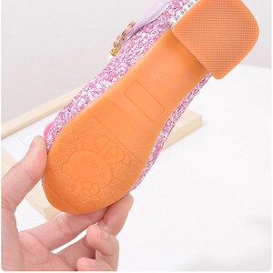 Puede incluir: Zapato rosa brillante para ni&ntilde;os con un tac&oacute;n peque&ntilde;o y una suela de goma. La suela es naranja y presenta un dise&ntilde;o de personaje de dibujos animados. El zapato tiene una hebilla y un detalle de lazo. El texto en la imagen dice "Tendon outsole".