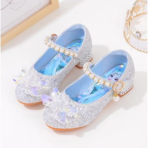 Puede incluir: Un par de zapatos plateados con purpurina estilo Mary Jane, con una correa de perlas y un tac&oacute;n peque&ntilde;o. Los zapatos tienen un interior azul claro con una imagen de personaje y una flor de cristal decorativa.