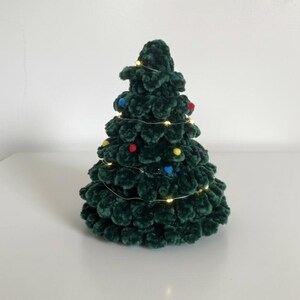 K&ouml;nnte beinhalten: Ein handgefertigter, dunkelgr&uuml;ner Weihnachtsbaum aus H&auml;kelarbeit mit bunten Pompons und Lichterketten. Der Baum ist eine festliche Dekoration f&uuml;r die Weihnachtszeit. Der Baum ist etwa 20 cm hoch.