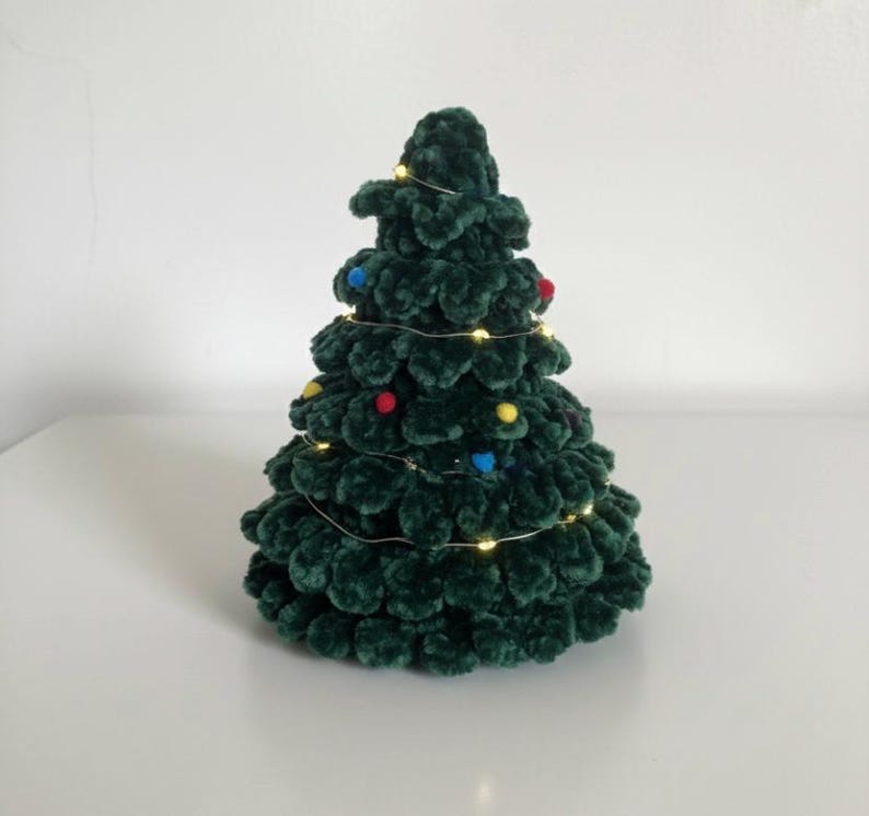 K&ouml;nnte beinhalten: Ein handgefertigter, dunkelgr&uuml;ner Weihnachtsbaum aus H&auml;kelarbeit mit bunten Pompons und Lichterketten. Der Baum ist eine festliche Dekoration f&uuml;r die Weihnachtszeit. Der Baum ist etwa 20 cm hoch.