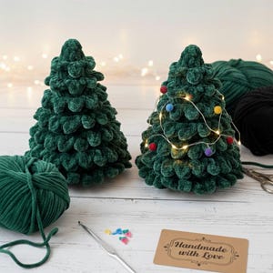 K&ouml;nnte beinhalten: Zwei handgefertigte gr&uuml;ne geh&auml;kelte Weihnachtsb&auml;ume. Einer ist mit einer Lichterkette und bunten Pompons geschm&uuml;ckt. Ein Kn&auml;uel gr&uuml;ne Wolle, eine H&auml;kelnadel und ein Etikett mit der Aufschrift "Handmade with Love" sind ebenfalls zu sehen.