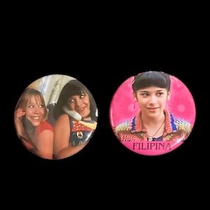 May include: Two round pink buttons with images of young girls. The first button shows two girls, one with a phone to her ear and the other with her hand on her ear. The second button shows a girl with a peace sign necklace and the text "FILIPINA".