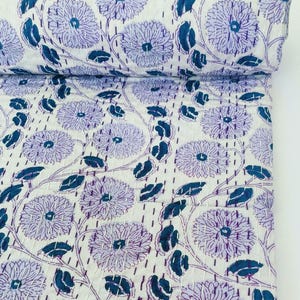 Op de afbeelding: Een witte katoenen quilt met een herhalend patroon van grote paarse bloemen en donkerblauwe bladeren. De stof is versierd met zichtbare rechte stiksels in een contrasterende draadkleur, wat zorgt voor een getextureerd uiterlijk over het oppervlak.