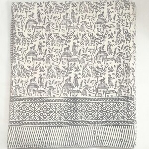 May include: A folded white textile featuring a repeating dark gray print of figures, animals, and huts. The bottom section displays a wide band with a geometric diamond pattern followed by a narrow border of diagonal stripes and visible stitching.