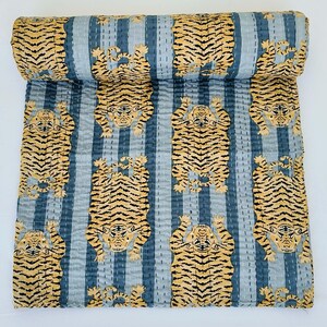 May include: A rolled-up, blue and white striped quilt featuring a repeating pattern of stylized tigers in shades of tan and black. The quilt is made of a soft, quilted fabric, and the tigers are arranged in vertical rows.