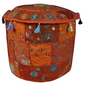 Op de afbeelding: Een ronde patchwork poef in tinten oranje, bruin en roest. De stof is versierd met geborduurde bloempatronen in blauw, groen en goud. Kwasten in blauw, geel en zwart hangen aan de bovenrand.