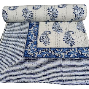 Op de afbeelding: Een opgerolde, gebroken witte en blauwe quilt met een paisley- en streepmotief. De quilt heeft een rand met bloemmotieven en een centraal gedeelte met paisley-motieven. Het kleurenschema is voornamelijk wit en blauw.