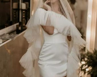 May include: A white satin mini dress with a ruffled tulle overlay. The dress is worn with a sheer veil and the model is looking at the camera.