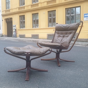 May include: A brown leather armchair and footstool set. The chair features a high back and a curved wooden frame. The footstool is low and has a similar design. The set is placed on a grey surface with a yellow building in the background.