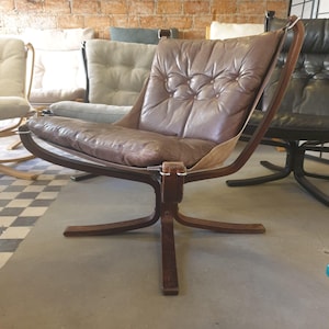May include: A vintage-style chair featuring a brown leather seat and a dark wooden frame. The chair has a distinctive design with a curved back and a four-legged base. The leather cushion is buttoned and shows signs of wear.
