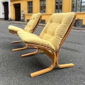 May include: Two mid-century modern lounge chairs with light yellow cushions and wooden frames. The chairs have a curved design and are placed on a gray asphalt surface. The background features a yellow building.