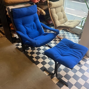Peut inclure: Un fauteuil bleu rembourré avec un repose-pieds assorti. Le fauteuil a un dossier haut et des accoudoirs, avec un cadre en métal noir. Le repose-pieds est rectangulaire. L'ensemble est posé sur un sol à carreaux noir et blanc.