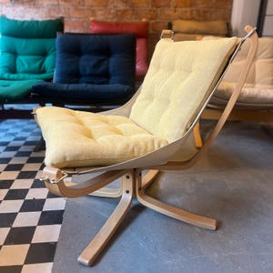 May include: A light yellow upholstered chair with a wooden frame and a canvas seat. The chair has a curved back and a padded seat cushion. Other chairs in various colours are visible in the background.