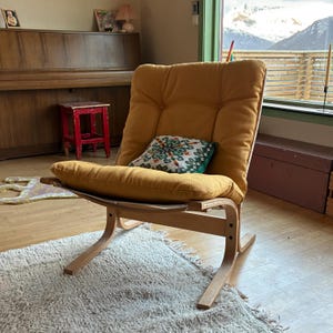 Könnte beinhalten: Ein Sessel im Mid-Century-Modern-Stil mit einem senfgelben Kissen und einem hellen Holzrahmen. Ein dekoratives Kissen mit grünem und weißem Muster liegt auf dem Sitz. Der Stuhl steht auf einem hellen Teppich.