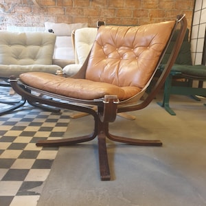 May include: A vintage-style chair featuring a brown leather seat and backrest. The chair has a dark wooden frame and a distinctive, curved design. The seat is buttoned, and the chair has a swivel base.