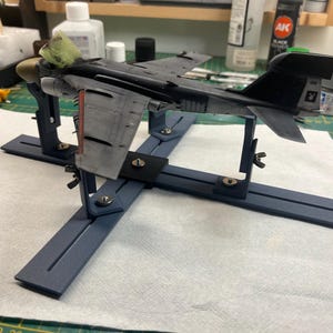Op de afbeelding: Een grijs en zwart modelvliegtuig is gemonteerd op een blauwe plastic modelbouwstandaard. Het vliegtuig wordt op zijn plaats gehouden door vier klemmen. De standaard heeft een kruisvorm met twee lange armen.