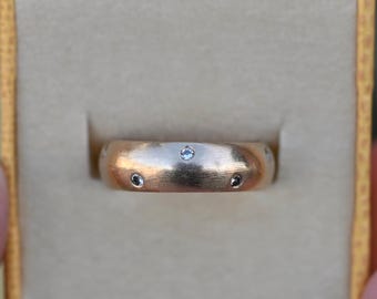 May include: A gold-colored ring with small, embedded gemstones is displayed inside a yellow ring box. The ring has a brushed metal finish and a rounded shape. The gemstones are evenly spaced around the band.