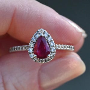 May include: A silver ring featuring a pear-cut ruby, surrounded by a halo of small diamonds. The ring's band is also set with small diamonds. The ruby is a rich red, and the diamonds sparkle. The ring is held in a hand.