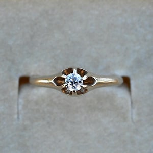 May include: A gold ring featuring a single round brilliant-cut clear gemstone set in a six-prong floral-shaped mounting. The band is smooth and tapers toward the center setting, which holds the stone securely in a polished yellow gold finish.