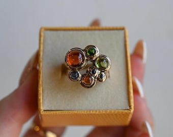 Könnte beinhalten: Ein goldener Ring mit einer Ansammlung farbiger Edelsteine. Der Ring zeigt einen großen orangefarbenen Edelstein, der von kleineren grünen, blauen und orangefarbenen Steinen umgeben ist. Der Ring wird in einer kleinen, quadratischen, goldfarbenen Schachtel präsentiert.