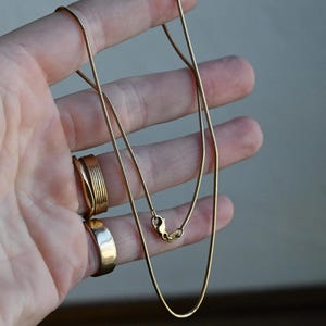 May include: A gold-toned snake chain necklace featuring a smooth, flexible link design and a secure lobster claw clasp. The chain has a polished finish and a consistent, rounded profile throughout its length, showcasing a classic and simple construction.
