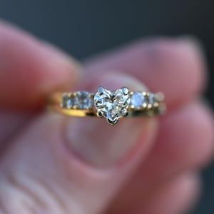 Può includere: Un anello d'oro con un diamante a forma di cuore come pietra centrale. La fascia è impreziosita anche da diamanti rotondi più piccoli. L'anello è presentato su uno sfondo sfocato, evidenziando la brillantezza delle gemme.