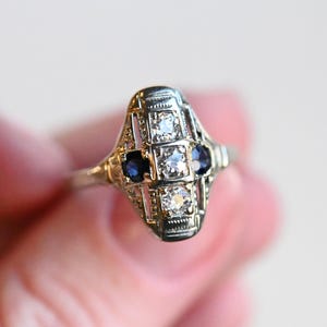 May include: A vintage ring with a delicate filigree design featuring three round gemstones. The center stone is a larger, clear diamond, flanked by two smaller, dark blue sapphires.