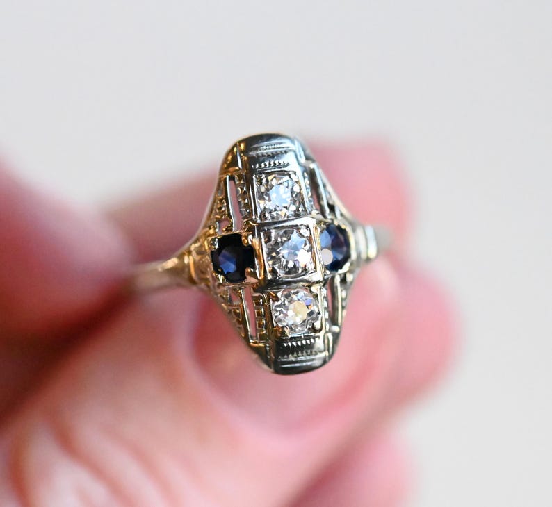 May include: A vintage ring with a delicate filigree design featuring three round gemstones. The center stone is a larger, clear diamond, flanked by two smaller, dark blue sapphires.