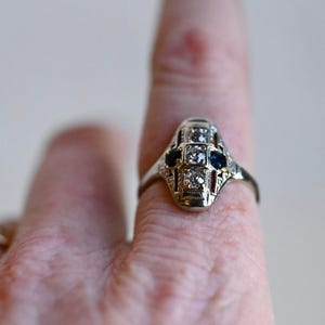 May include: A silver ring with a geometric design featuring a central diamond and two blue sapphires.