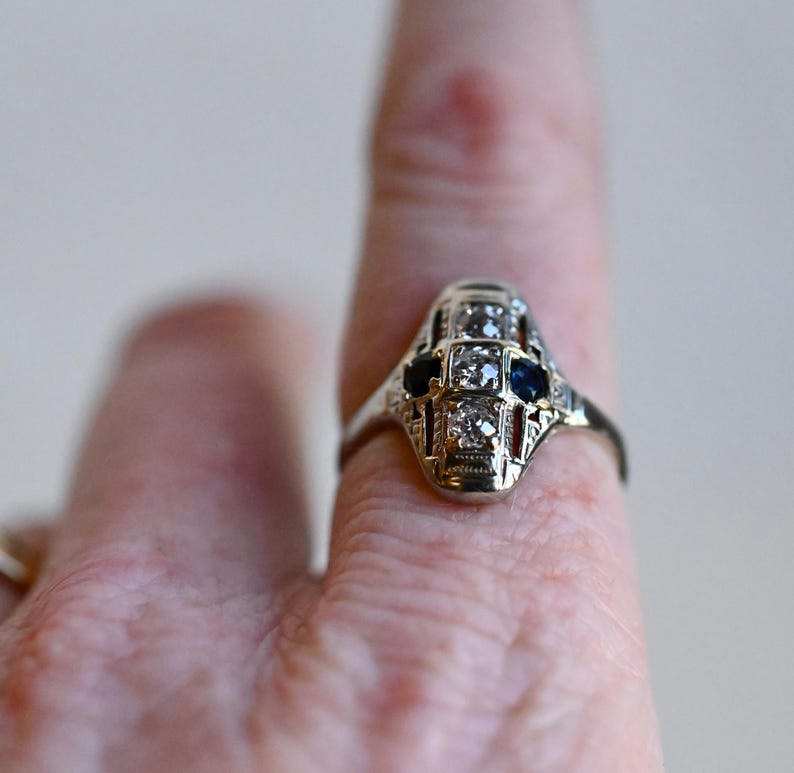 May include: A silver ring with a geometric design featuring a central diamond and two blue sapphires.