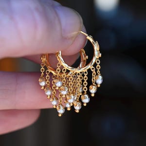 Puede incluir: Un par de pendientes de aro dorados con múltiples cadenas colgantes, cada una rematada con una pequeña perla blanca. Los aros circulares tienen un acabado pulido y la franja de cadenas en cascada crea una apariencia texturizada y en capas.