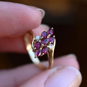May include: A gold ring featuring a cluster of six marquise-cut deep red rubies arranged in two vertical rows, accented by two small round-cut clear diamonds positioned at the top and bottom of the ruby setting on a polished gold band.