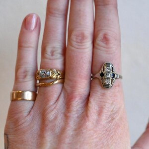 May include: Two rings on a hand. The ring on the index finger is a silver band with a square-shaped center stone and two smaller stones on either side. The ring on the middle finger is a gold band with a leaf design.