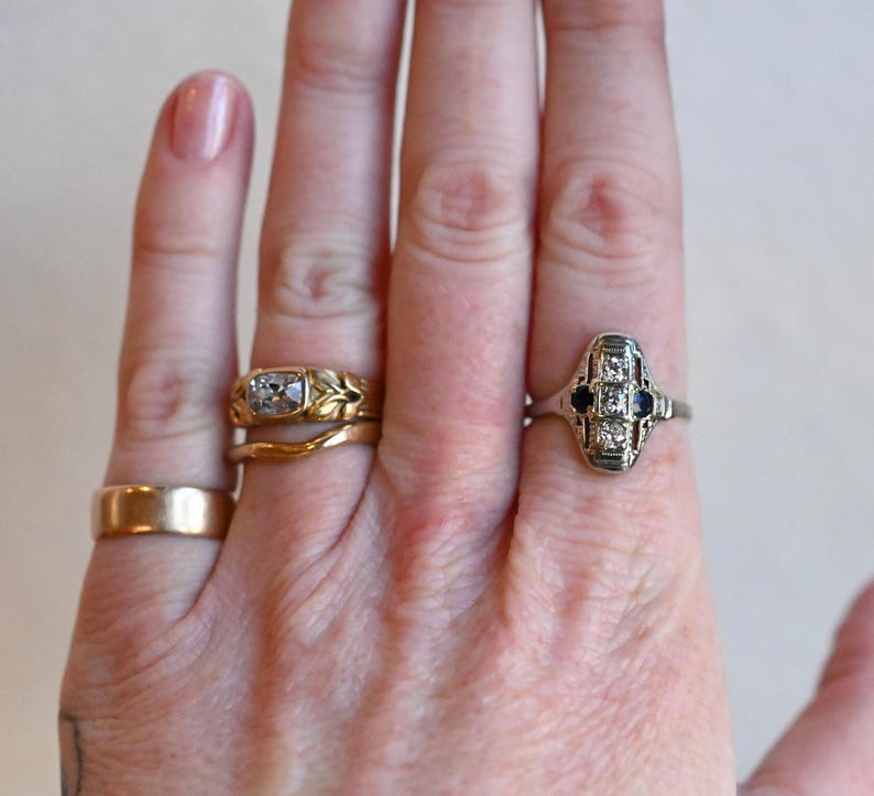 May include: Two rings on a hand. The ring on the index finger is a silver band with a square-shaped center stone and two smaller stones on either side. The ring on the middle finger is a gold band with a leaf design.