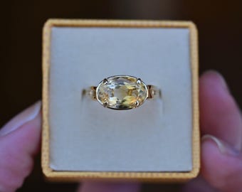 May include: A gold ring with a large, oval-cut yellow gemstone. The ring is set in a gold band and is presented in a small, beige jewelry box. The ring is a piece of jewelry.