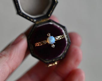 May include: A gold ring with a central, round, light blue opal gemstone, accented by small diamonds. The ring is displayed in a vintage, octagonal, dark blue jewelry box with a purple velvet interior. The ring's band features delicate, ornate detailing.