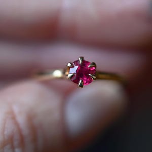 Puede incluir: Un anillo de oro con una piedra preciosa redonda, facetada y de color rojo rosado intenso, montada en un engaste de seis garras. La banda es lisa y metálica, con un acabado pulido que refleja la luz. La piedra es el punto central de la pieza.