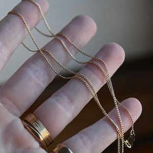 May include: A thin, gold-colored metal chain necklace featuring small, interlocking links. The delicate chain is draped across fingers, showcasing its fine texture and reflective surface. A small, flat clasp is visible at the end of the chain.