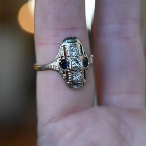 May include: A vintage ring with a geometric design featuring a central diamond and two blue sapphires. The ring is set in white gold and has a delicate filigree pattern.