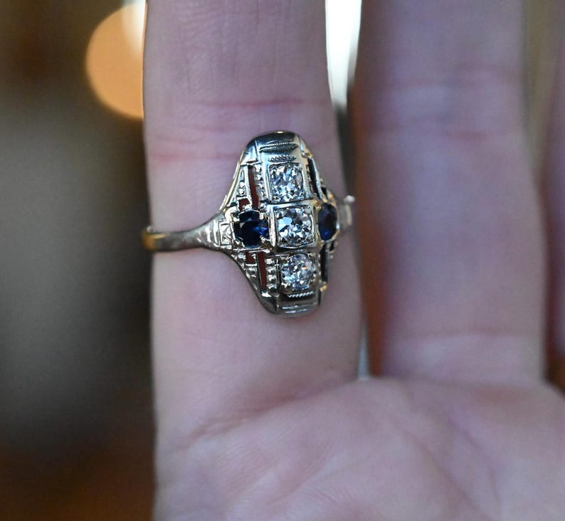 May include: A vintage ring with a geometric design featuring a central diamond and two blue sapphires. The ring is set in white gold and has a delicate filigree pattern.