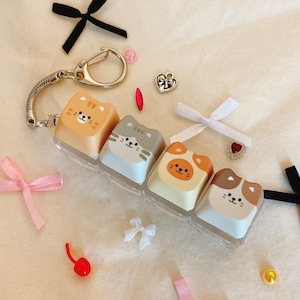 May include: A keychain featuring four cat-shaped keycaps in orange, gray, and brown. The keycaps have cat faces and are attached to a silver chain with a clasp. The image also includes small bows and decorative charms.